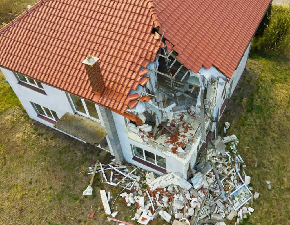 Aerial view of a damaged house with a red tiled roof in Palm Beach County. The corner structure has collapsed, scattering debris on the ground. The remaining roof and walls are intact, surrounded by a grassy area. This site is ideal for storm damage restoration services.