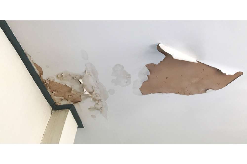 An image of a ceiling with significant water damage, showing peeling paint, damp patches, and brown stains. The damage is concentrated near a corner, with a piece of paint hanging down.