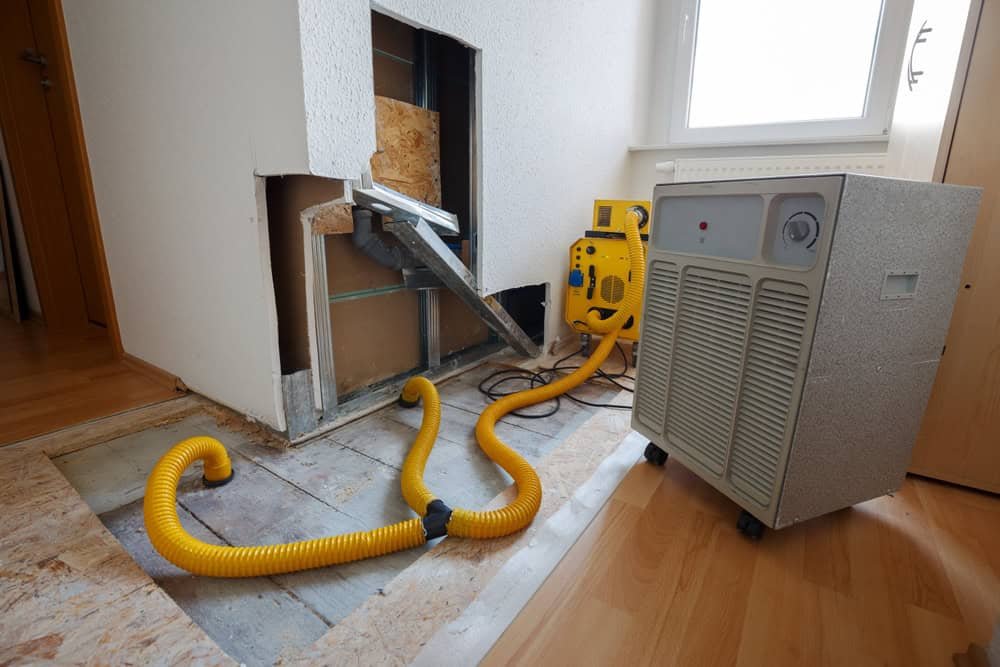 A room with a partially open wall and missing flooring. A yellow hose is attached to a dehumidifier, and another yellow machine is nearby. The setup suggests a drying or restoration process, likely due to water damage.