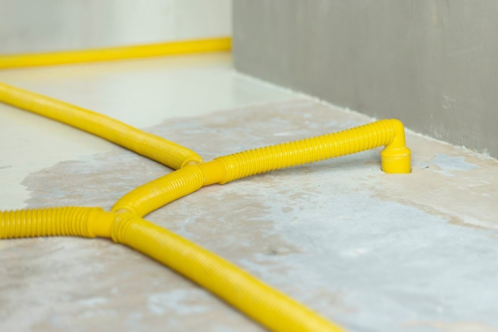 Yellow flexible ducts lie across a bare floor, connecting to a wall. The floor appears unfinished, with a mix of gray and cement textures. The ducts have a corrugated texture and extend into the corner.